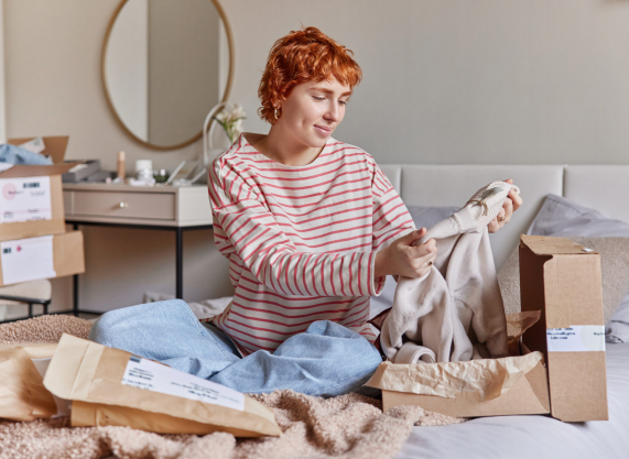 Young happy redhead woman wears striped jumper and jeans packs purchase for selling poses on comfortable bed against bedroom interior gets her online order checks delivery. Shopping from home